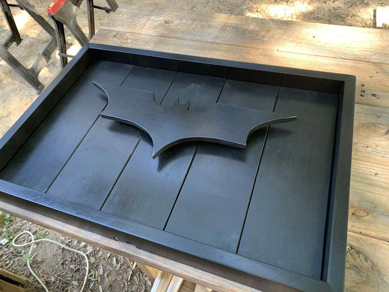 May include: A rectangular black wooden tray featuring a raised, matte black Batman bat-symbol emblem centered on the base. The tray is constructed from vertical wooden planks with a smooth, dark finish and a solid frame border surrounding the interior.