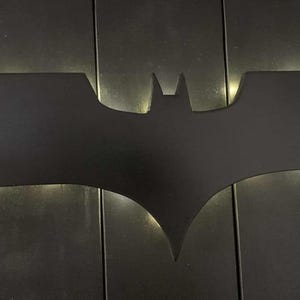 May include: A matte black wall-mounted light fixture shaped like the iconic Batman bat symbol. The silhouette features sharp, angular wings and a central head section, with a soft white glow emanating from behind the edges to create a halo effect.