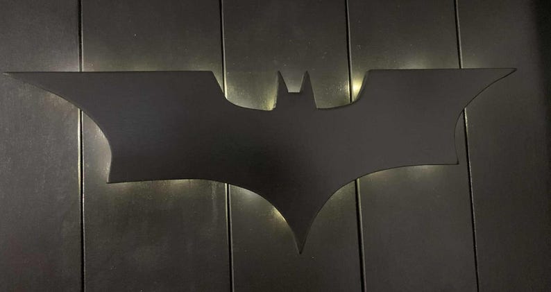 May include: A matte black wall-mounted light fixture shaped like the iconic Batman bat symbol. The silhouette features sharp, angular wings and a central head section, with a soft white glow emanating from behind the edges to create a halo effect.