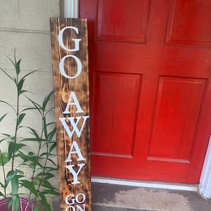 May include: A wooden sign with the words "GO AWAY GO ON GIT" in white lettering. The sign has a dark brown, burnt wood finish and is leaning against a red door. A potted plant is to the left. The sign is likely for a front door.
