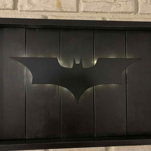 May include: A rectangular black wall art piece featuring a centered, cutout Batman bat symbol. The design is composed of vertical black panels set within a deep black frame, with soft white light emanating from behind the bat silhouette.