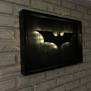 May include: A rectangular black wooden wall sign featuring a cutout of the Batman bat symbol in the center. The sign is illuminated from behind the symbol, casting a warm glow against the dark vertical wooden planks of the frame's interior.