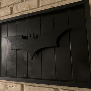 May include: A rectangular black wooden wall sign featuring a raised, centered bat-shaped silhouette. The background consists of vertical wooden planks framed by a solid black border, creating a monochromatic, textured appearance for this wall decor.