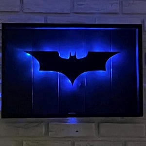 May include: A rectangular wall-mounted sign featuring a black silhouette of a bat emblem centered on a dark background. The sign is illuminated from behind by a vibrant blue LED light that creates a glowing outline around the bat shape.