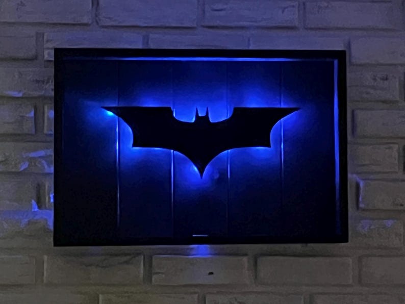 May include: A rectangular wall-mounted sign featuring a black silhouette of a bat emblem centered on a dark background. The sign is illuminated from behind by a vibrant blue LED light that creates a glowing outline around the bat shape.