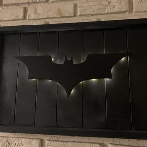 May include: A rectangular wall sign featuring a black wooden frame and a black vertical plank background. A black bat-shaped silhouette is mounted in the center, illuminated from behind by soft white light that creates a glowing outline effect.