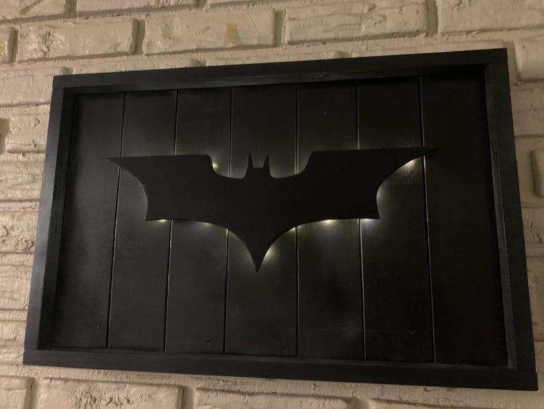 May include: A rectangular wall sign featuring a black wooden frame and a black vertical plank background. A black bat-shaped silhouette is mounted in the center, illuminated from behind by soft white light that creates a glowing outline effect.