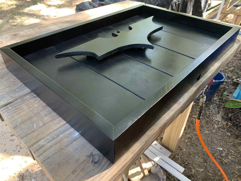 May include: A rectangular black wooden tray featuring a raised black bat-shaped emblem centered on the base. The tray has a smooth, matte finish with clean, straight edges and a slatted bottom design, providing a flat surface for display or storage.
