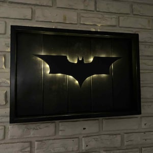 May include: A rectangular wall-mounted sign featuring a black Batman logo silhouette centered on a dark, vertical-slatted wooden panel. The logo is backlit with a soft, warm glow that illuminates the edges, all enclosed within a black frame.