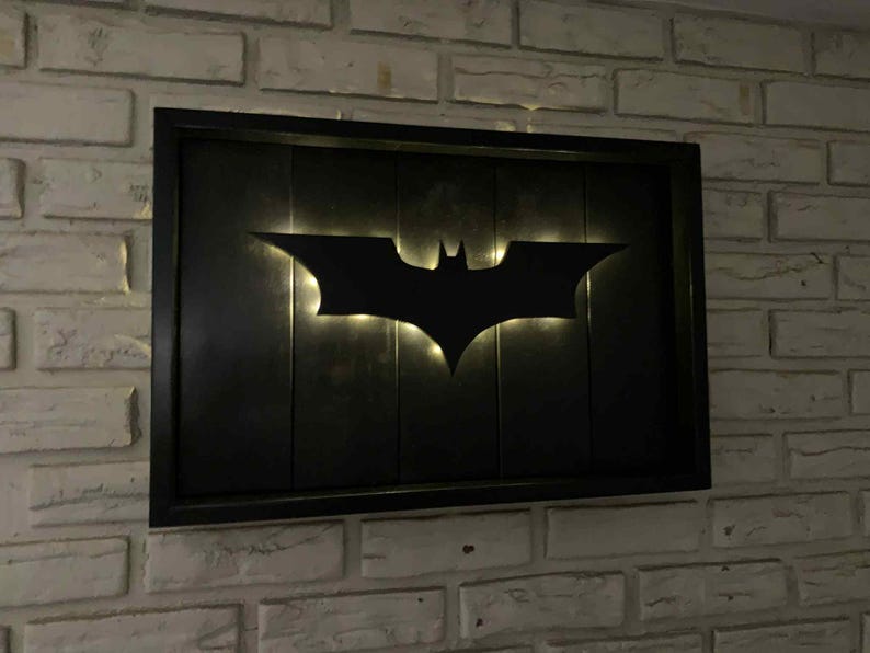 May include: A rectangular wall-mounted sign featuring a black Batman logo silhouette centered on a dark, vertical-slatted wooden panel. The logo is backlit with a soft, warm glow that illuminates the edges, all enclosed within a black frame.