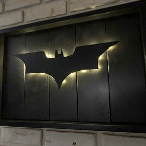 May include: A rectangular wall-mounted sign featuring a black wooden frame and a black vertical plank background. A matte black Batman bat symbol is mounted in the center, illuminated from behind by warm white LED lights that create a glowing effect.