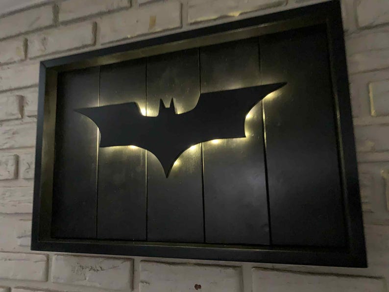 May include: A rectangular wall-mounted sign featuring a black wooden frame and a black vertical plank background. A matte black Batman bat symbol is mounted in the center, illuminated from behind by warm white LED lights that create a glowing effect.