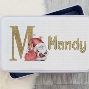 Puede incluir: Una lata rectangular con fondo blanco y una "M" dorada con purpurina y el nombre "Mandy" en dorado. Un gnomo de Papá Noel con gorro rojo y saco rojo está en el lado derecho de la lata. La lata está abierta, revelando un interior azul oscuro.