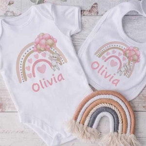 May include: A white baby bodysuit and bib with a pink and brown rainbow design, a bunny holding balloons, and the name "Olivia" printed on both. A matching rainbow-shaped woven wall hanging is also included.