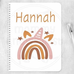 Könnte beinhalten: Ein weißes Notizbuch mit Spiralbindung. Der Einband zeigt einen rosa, orangefarbenen und braunen Regenbogen mit einem Einhornhorn und Sternen. Der Name "Hannah" ist oben auf dem Notizbuch in Orange gedruckt.