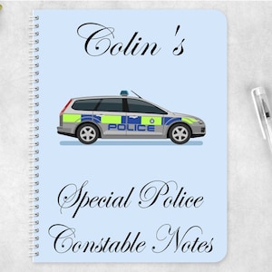 Puede incluir: Una libreta azul con una encuadernación en espiral blanca. La portada presenta un coche de policía de dibujos animados con la palabra "POLICE" en el lateral. El texto "Colin's Special Police Constable Notes" está escrito en cursiva en la portada.