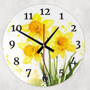 May include: A white round clock with black hands and black numbers. The clock face features a watercolour painting of yellow daffodils with green leaves on a white background.