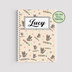 Può includere: Un quaderno a spirale color crema con un motivo di gatti grigi dei cartoni animati, impronte di zampe e la parola "MEOW". Il nome "Lucy" è visualizzato in un rettangolo bianco. Il quaderno è A5/A4 Eco-Friendly.