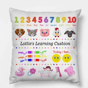 Könnte beinhalten: Weißes Kissen mit bunten Zahlen, Tiergesichtern und dem Text "Lottie's Learning Cushion". Enthält Buntstifte, Smileys und andere Illustrationen. Ein lustiges und lehrreiches Kissen für Kinder.