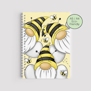 May include: A spiral-bound notebook with a whimsical design featuring bee-themed gnomes. The gnomes have yellow and black striped hats and white beards, set against a light yellow background with small bee illustrations. The notebook has a green circle with the text "A5 / A4 Eco-Friendly".