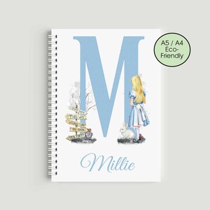 May include: A white spiral-bound notebook with a blue polka-dot letter "M" on the cover. The notebook features an illustration of Alice in Wonderland, a white rabbit, and a signpost. The name "Millie" is written in cursive script.