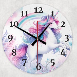Może przedstawiać: Biały jednorożec z różową i fioletową grzywą i ogonem stoi na białym tle z tęczą i kwiatowymi akcentami. Jednorożec ma złoty róg i zamknięte oczy. Tarcza zegara ma czarne rzymskie cyfry godzinowe i czarne wskazówki.