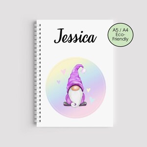 May include: A spiral-bound notebook with the name "Jessica" in black script. The cover features a whimsical illustration of a purple gnome with a white beard, set against a pastel rainbow background. A green circle indicates the notebook is A5/A4 and eco-friendly.