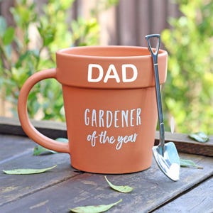 Può includere: Tazza in ceramica color terracotta a forma di vaso, con la parola "DAD" in lettere bianche in alto. Sotto, il testo recita "GARDENER of the year". Una piccola cazzuola d'argento è appoggiata accanto alla tazza.