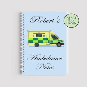 Może przedstawiać: Spiralny notatnik w kolorze jasnoniebieskim z ilustracją żółto-zielonej karetki. Tekst "Robert's Ambulance Notes" jest napisany elegancką czcionką. Zielone kółko wskazuje, że notatnik jest A5/A4 Eco-Friendly.