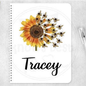 Può includere: Un quaderno bianco con rilegatura a spirale presenta un girasole con api che volano intorno. Il nome "Tracey" è scritto in corsivo nero nella parte inferiore del quaderno.