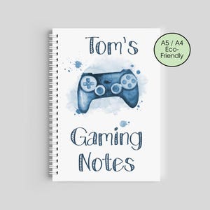 Puede incluir: Un cuaderno de espiral blanco con un diseño de acuarela azul de un mando de videojuegos. El texto "Tom's Gaming Notes" está escrito encima y debajo del mando. Un círculo verde indica que el cuaderno es A5/A4 y ecológico.