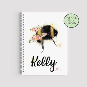 Może przedstawiać: Biały notatnik w spiralę z akwarelową ilustracją trzmiela ozdobionego różowymi kwiatami. Imię "Kelly" jest napisane kursywą pod pszczołą. Zielone kółko wskazuje, że notatnik jest A5/A4 i ekologiczny.
