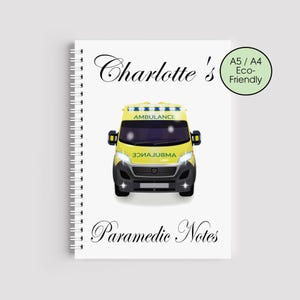 Puede incluir: Un cuaderno encuadernado en espiral blanco con una ilustración de ambulancia amarilla. El cuaderno tiene el texto "Charlotte's" y "Paramedic Notes". Un círculo verde indica que es A5/A4 y ecológico.