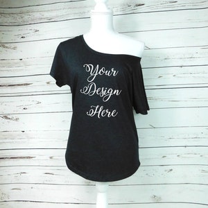 May include: A black off-the-shoulder t-shirt with the text "Your Design Here" printed in white on the front.