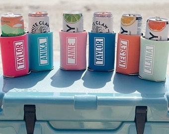 Puede incluir: Seis enfriadores de latas personalizados en varios colores, incluyendo rosa, azul, verde y naranja, con nombres impresos en cada enfriador. Los enfriadores están colocados encima de una nevera Yeti azul con el logotipo de Yeti y las palabras "Chessa Creations" impresas en el lateral.