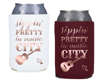 Puede incluir: Dos enfriadores de latas, uno blanco y otro marrón, con el texto "sippin' pretty in music city" en letras doradas rosas. Una guitarra rosa dorada está representada en cada enfriador.