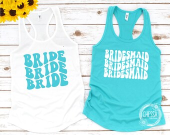 Op de afbeelding: Twee witte tanktops met een turquoise ontwerp. Het topje aan de linkerkant zegt "BRIDE" drie keer in een retro-lettertype. Het topje aan de rechterkant zegt "BRIDESMAID" drie keer in hetzelfde lettertype.