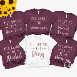 Puede incluir: Seis camisetas marrones y blancas con texto dorado rosado. Las camisetas dicen "I'll bring the" seguido de "Dance Moves", "Shenanigans", "Voice of Reason", "Alcohol", "Ring" y "Bad Decisions".