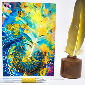 May include: An art print with a feather design, gold leaf accents, a brown inkwell with a gold feather, and a yellow pen. The artwork has a vibrant mix of blue, yellow, and gold colours. The inkwell is approximately 5 cm tall.