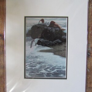 May include: A framed print of a woman lying on a rock by the ocean. The woman is wearing a white shirt and dark pants. A seagull flies in the background.