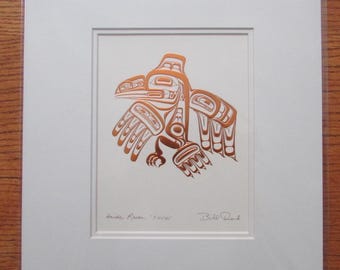 May include: A framed print featuring a stylized copper-colored raven design. The artwork, set against a white mat, showcases intricate details and is signed by the artist. The print is labeled "Haida Raven 'Xuuya'" and includes a "Made in Canada" sticker.