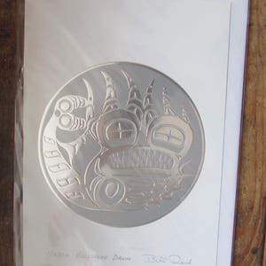 May include: A silver print of a Haida Bullhead Drum design. The artwork features a circular design with intricate patterns and shapes, including a stylized face and other symbolic elements. The print is on a white background.