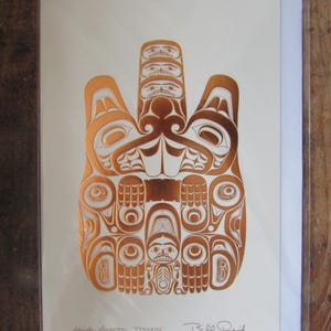 Puede incluir: Una impresión de color cobre de un diseño de castor Haida sobre papel color crema. La obra de arte presenta patrones y figuras intrincados, con la firma del artista en la esquina inferior derecha. La impresión está dentro de una funda de plástico transparente.