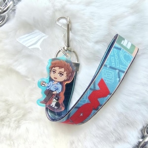 May include: A keychain featuring a cartoon illustration of a person and a cat. The keychain has a blue and red lanyard with a silver clasp. The illustration is on a clear acrylic charm.