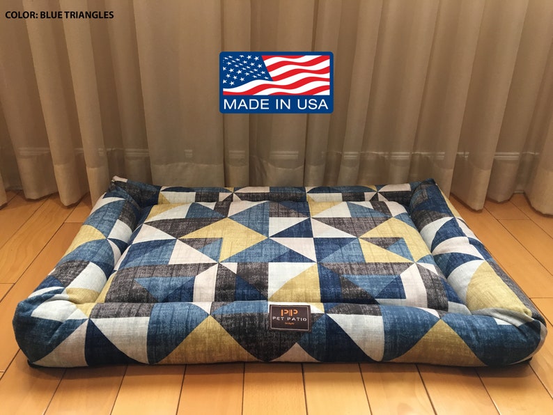 May include: A rectangular pet bed with a blue, yellow, and white geometric pattern. The bed has a raised edge and is made in the USA. The text "PIP PET PATIO" is visible on the bed.