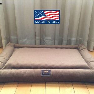 May include: A brown rectangular dog bed with a raised edge and a brown fabric label that says "P.P. PET PATIO" in gold lettering. The bed is on a wooden floor in front of a window with sheer curtains.