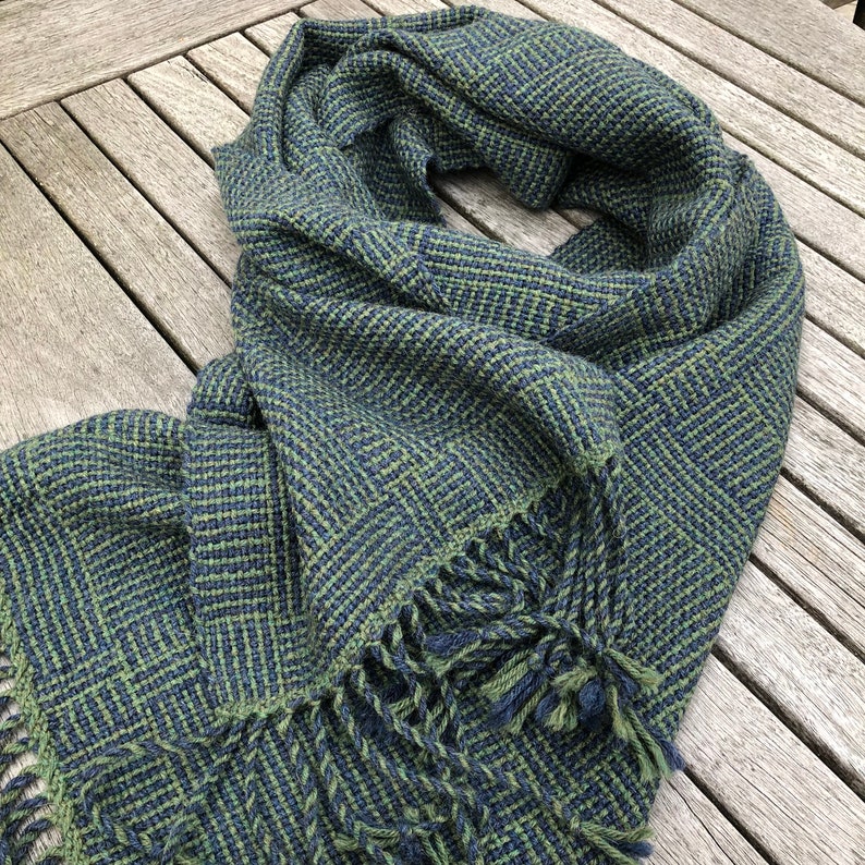 K&ouml;nnte beinhalten: Ein gr&uuml;n-blauer gewebter Schal mit geometrischem Muster und Fransen.