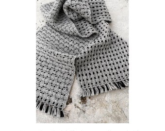 Könnte beinhalten: Grau-schwarzer gewebter Schal mit strukturiertem Muster und Fransen. Der Schal trägt die Aufschrift "Ripples & Waves Scarf" von Christine Jablonski. Der Schal ist aus Alpaka gefertigt und ein Winter-Basic.