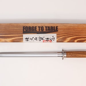 May include: A silver steel knife sharpener with a wooden handle and a wooden box with the text "FORGE TO TABLE" on the top.