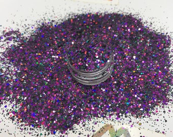 Op de afbeelding: Een close-up van een hoop paarse en zwarte glitter met iriserende vlekken. Een kleine, doorzichtige container is gevuld met de glitter en staat in het midden van de hoop. De glitter is verspreid op een wit oppervlak.